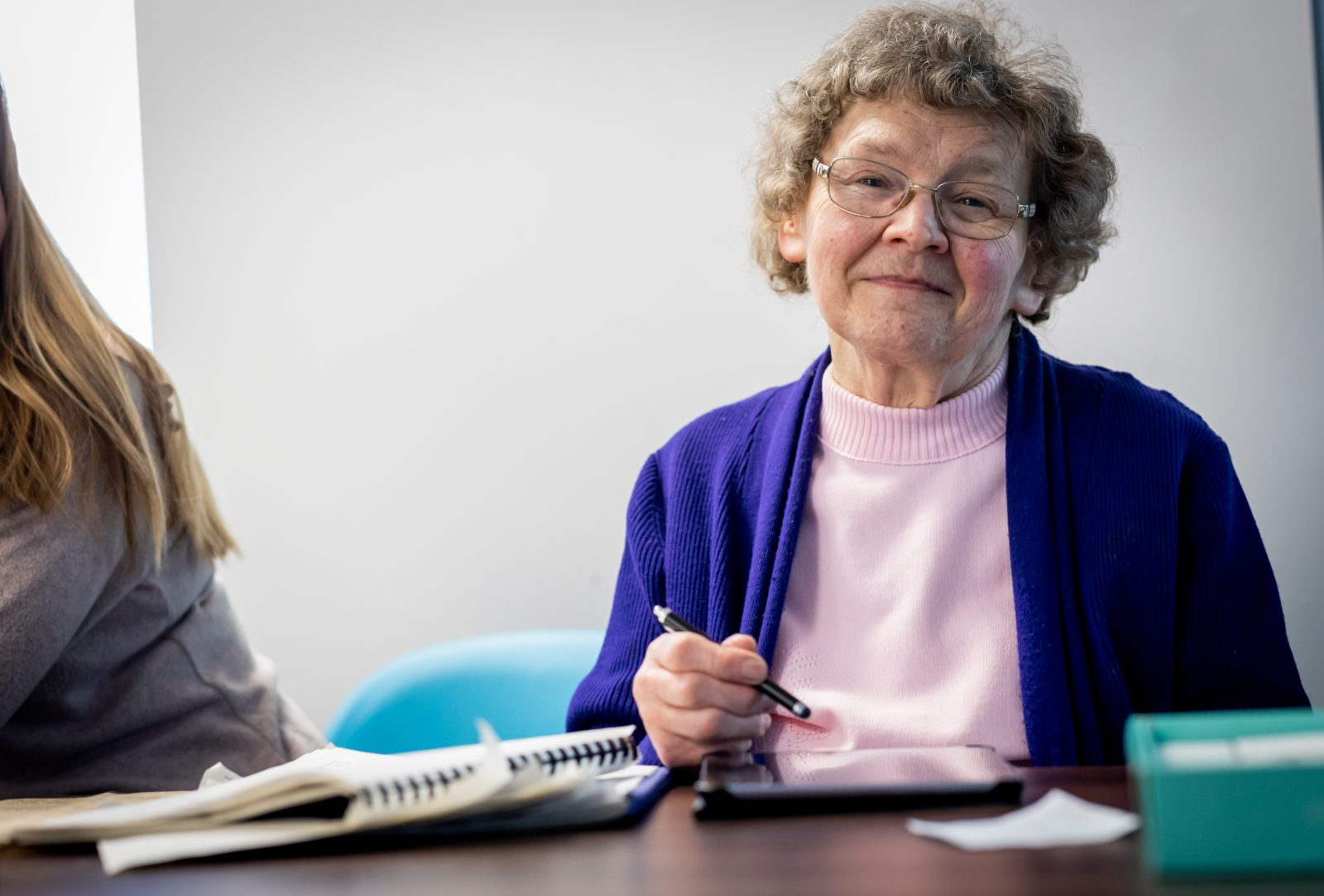 An older woman in a pink top and purple cardigan smiling while going through paperwork at a table with a helper beside her.