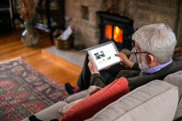 An older man relaxing on the sofa by the fire, reading on his tablet.