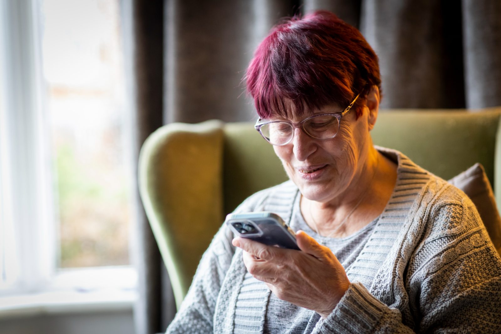 An older woman sitting in an armchair reading a reminder aloud from her smartphone.